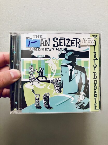 Hand holding a CD jewel case for The Brian Setzer Orchestra’s The Dirty Boogie, featuring retro teal, black, and green swing-era artwork. Price stickers on the cover read “1-” and “Lyle’s Place $12.95.”