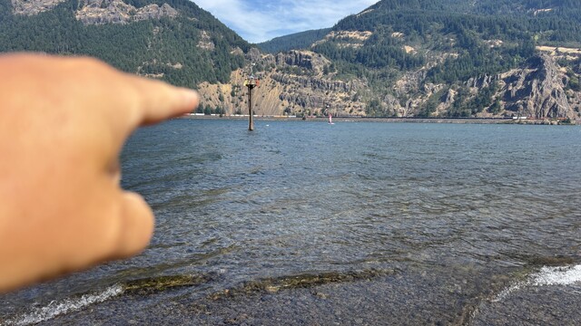 A finger points towards a distant pole in the water, with a backdrop of green hills and rocky cliffs. The foreground features clear water and small waves, while the background includes a glimpse of a shoreline and a few boats.