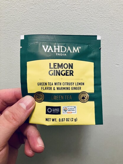 A hand holds a single Vahdam India Lemon Ginger green tea packet against a light textured wall. The dark green and yellow sachet reads “Green Tea with Citrusy Lemon Flavor & Warming Ginger,” and contains a 2 g pyramid tea bag.